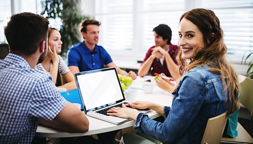 Una chica sonrie y voltea a la cámara mientras está sentada en una mesa de trabajo frente a una computadora, mientras otros de sus compañeros conversan en la misma mesa