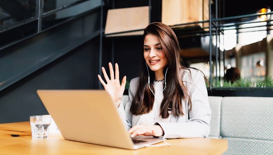 Una chica sonríe a cámara y saluda con la mano, en un escritorio tras una computadora